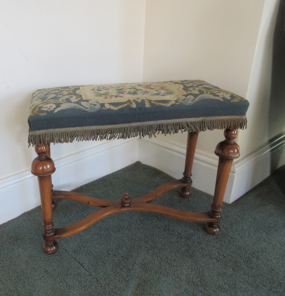 An antique Victorian walnut stool features an X-frame stretcher base and a rectangular seat upholstered in patterned blue needlepoint with fringing.