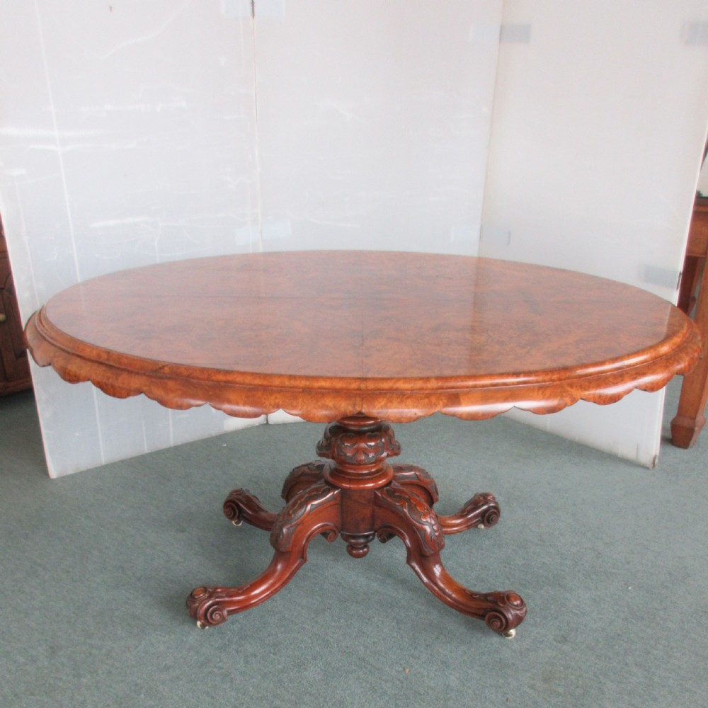 An antique Victorian oval tilt-top loo table made of figured burr walnut with a carved pedestal, four carved legs, and brass castors.