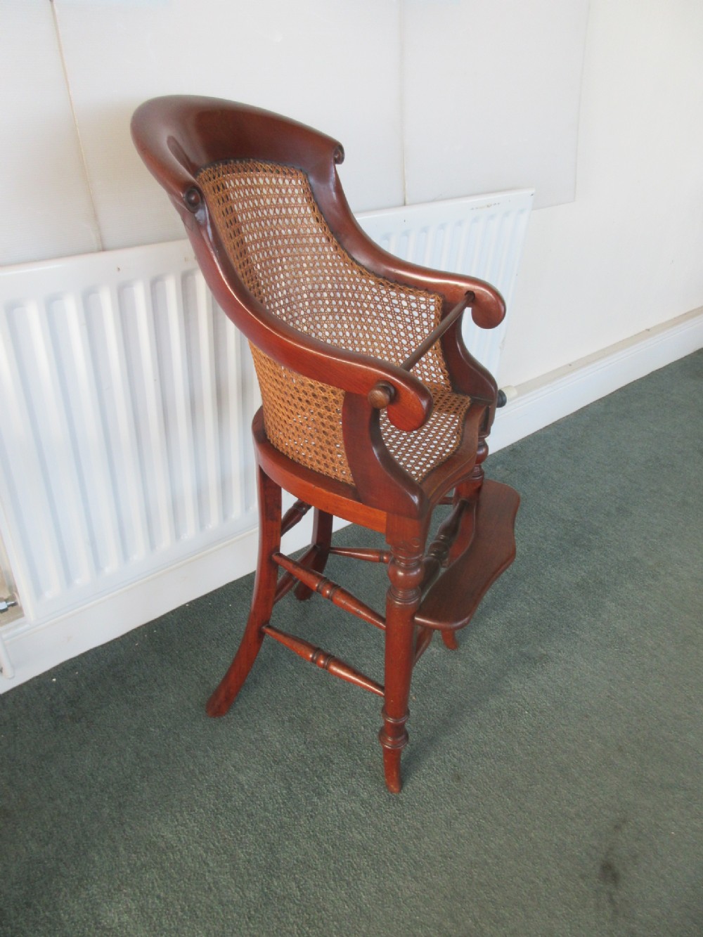 A Victorian mahogany childs high chair with a woven cane backrest, curved armrests, a safety bar, turned legs, and an integrated wooden footrest.
