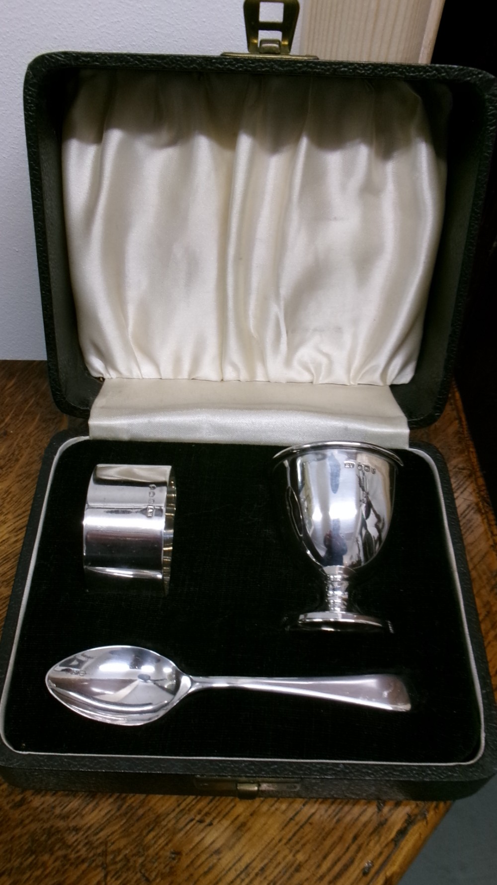 A three-piece sterling silver christening set comprising an egg cup, napkin ring, and spoon, presented inside a green velvet and silk-lined box.