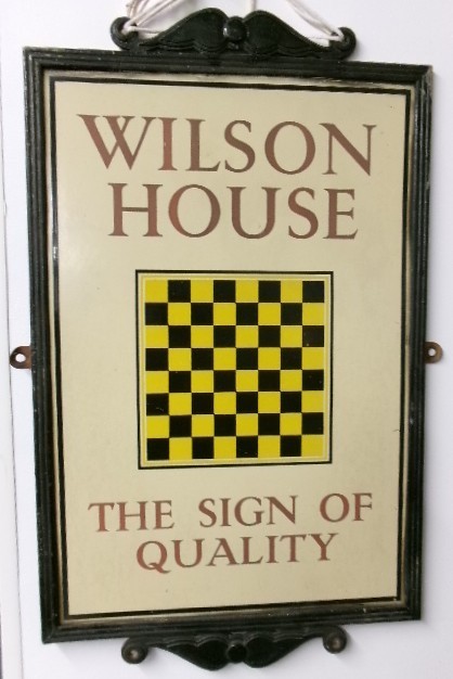 A rectangular framed cream enamel advertising sign displaying the text Wilson House The Sign of Quality around a central yellow and black chequered square. View V