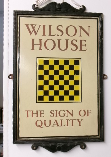 A rectangular framed cream enamel advertising sign displaying the text Wilson House The Sign of Quality around a central yellow and black chequered square.