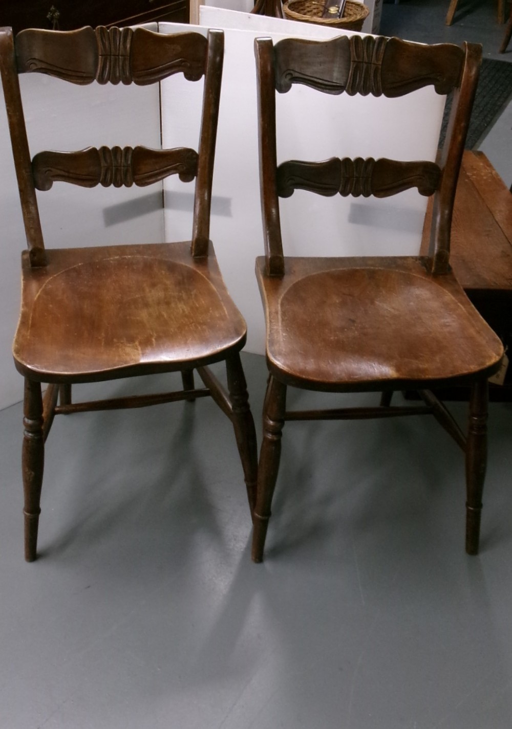 A pair of late Victorian dark brown wooden kitchen chairs with carved back rails, saddled wooden seats, and turned front legs.