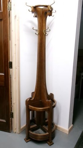 A tall solid oak hat and coat stand featuring multiple brass hooks, a tapered central column, and an integrated circular umbrella holder base.
