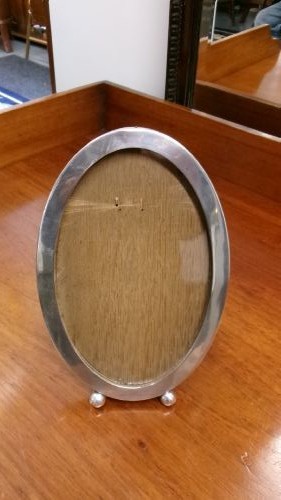 An oval sterling silver photo frame with a plain border, two ball feet, and a wooden backing.