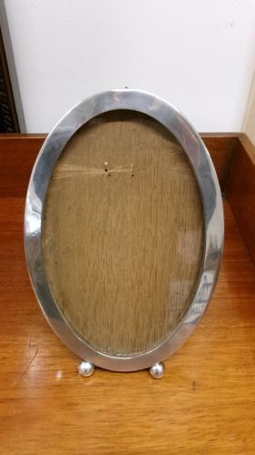 An oval sterling silver photo frame with a plain border, two ball feet, and a wooden backing. View V
