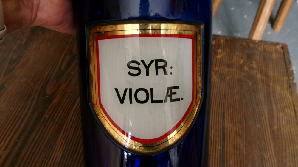 A tall Victorian cobalt blue glass apothecary bottle featuring a dark glass lollipop stopper and a gilt-bordered shield label inscribed SYR VIOLAE. View VIII