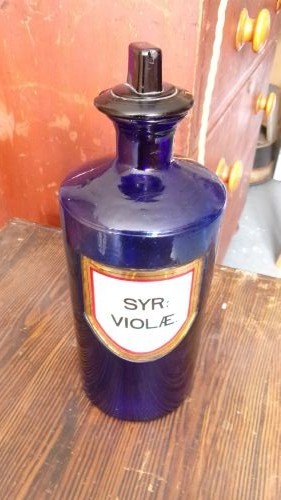 A tall Victorian cobalt blue glass apothecary bottle featuring a dark glass lollipop stopper and a gilt-bordered shield label inscribed SYR VIOLAE.