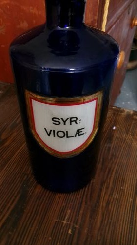 A tall Victorian cobalt blue glass apothecary bottle featuring a dark glass lollipop stopper and a gilt-bordered shield label inscribed SYR VIOLAE. View III