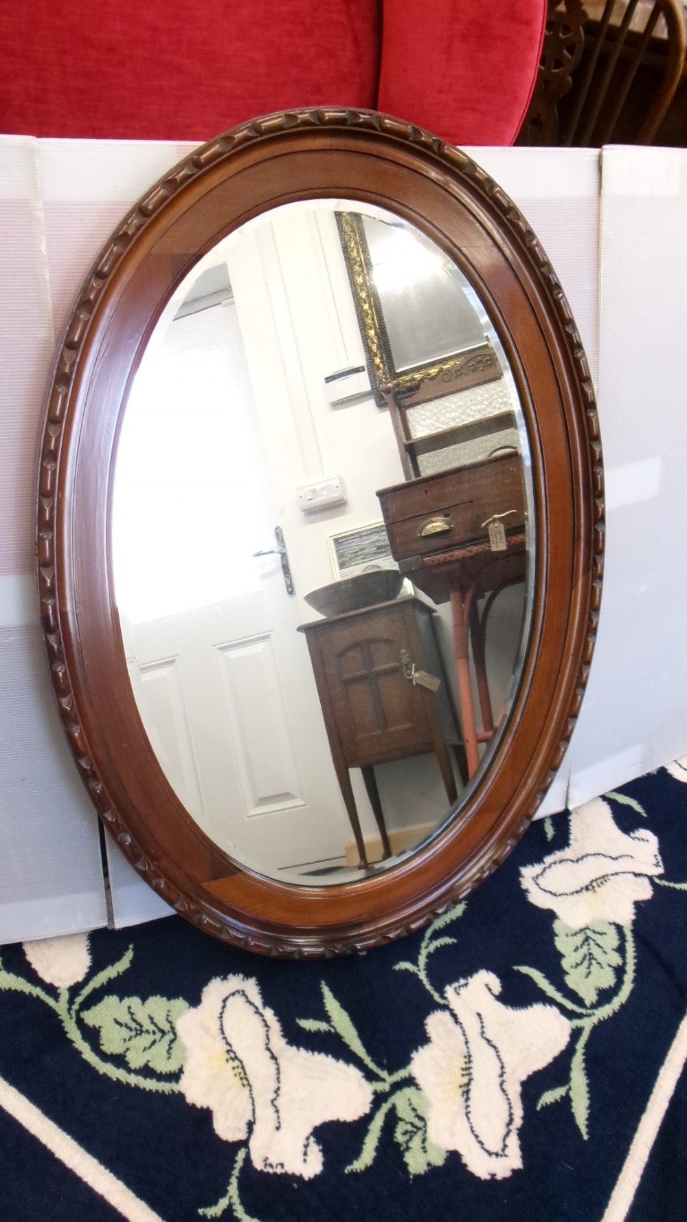 An Edwardian oval wall mirror crafted from mahogany timber featuring a carved decorative outer rim and bevelled glass.
