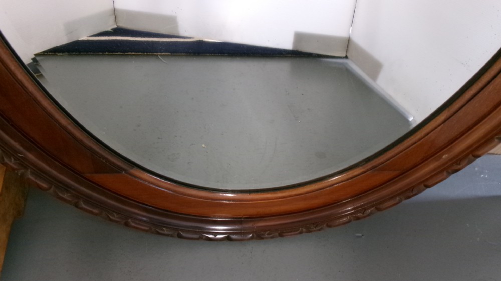 An Edwardian oval wall mirror crafted from mahogany timber featuring a carved decorative outer rim and bevelled glass. View IV