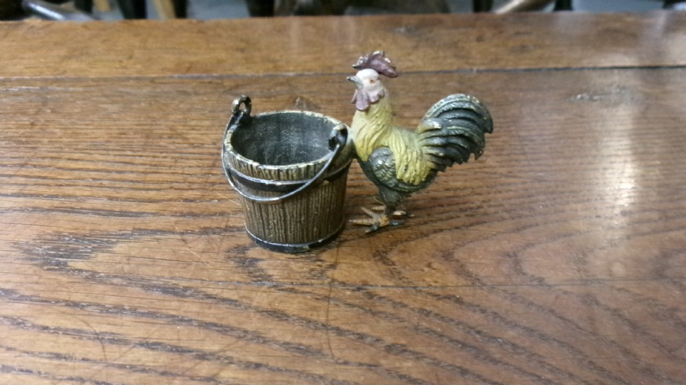 A small cold-painted bronze figurine depicting a cockerel standing alongside a miniature metal bucket with a handle.