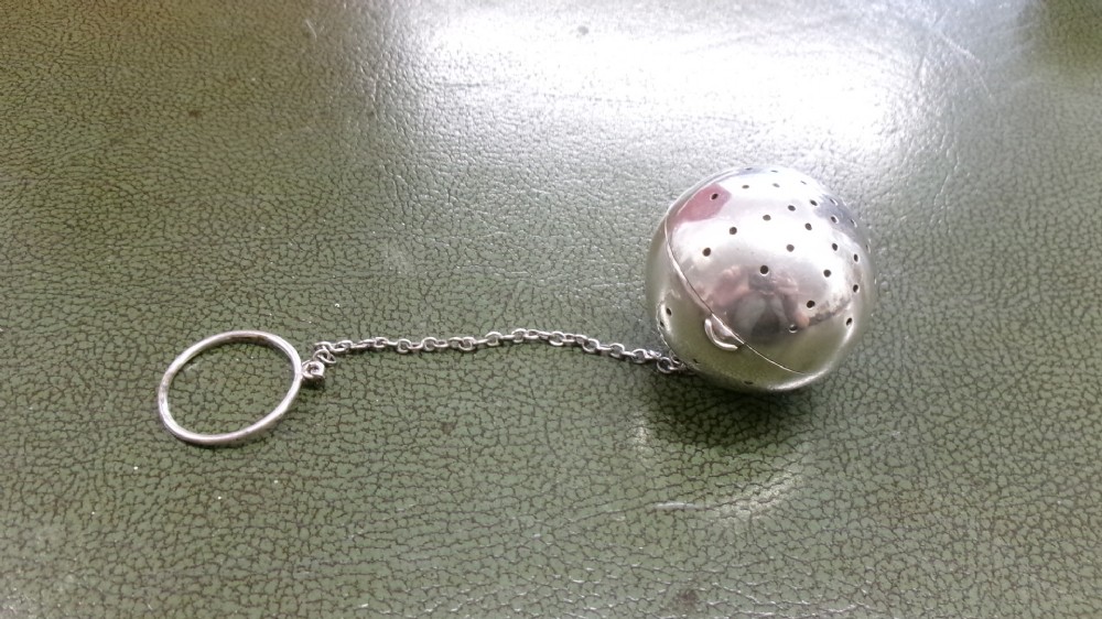 A spherical sterling silver tea infuser featuring fine round perforations, a hinged opening, and a chain attached to a finger ring.