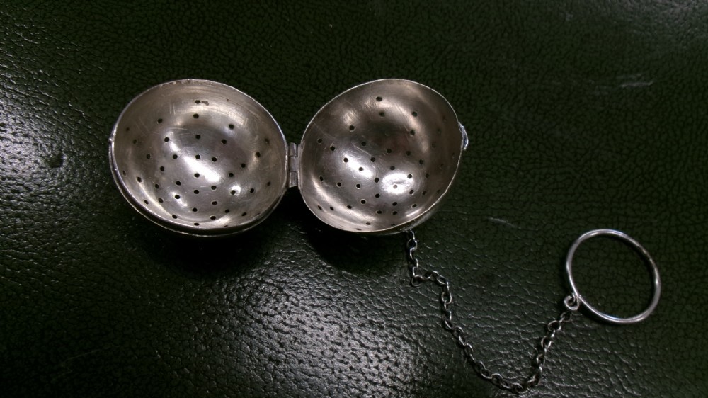 A spherical sterling silver tea infuser featuring fine round perforations, a hinged opening, and a chain attached to a finger ring. View II