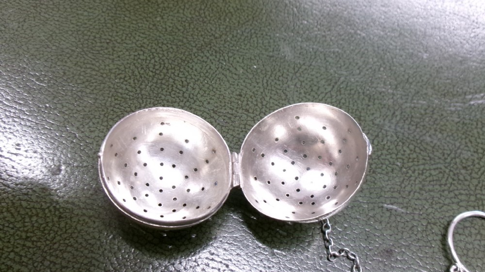 A spherical sterling silver tea infuser featuring fine round perforations, a hinged opening, and a chain attached to a finger ring. View III