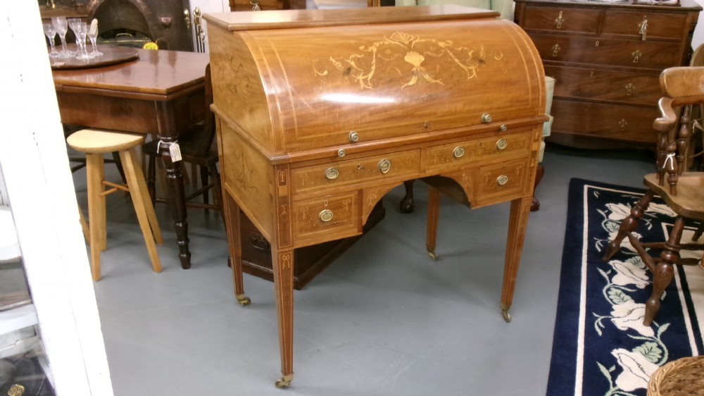 An antique Edwardian inlaid mahogany cylinder top writing desk featuring an open curved roll top, fitted internal compartments, a green leather writing surface, and tapered square legs with brass castors. View II