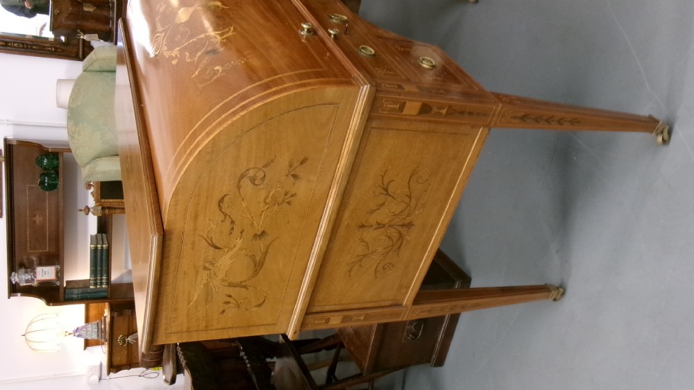 An antique Edwardian inlaid mahogany cylinder top writing desk featuring an open curved roll top, fitted internal compartments, a green leather writing surface, and tapered square legs with brass castors. View III