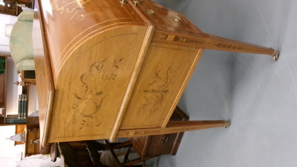An antique Edwardian inlaid mahogany cylinder top writing desk featuring an open curved roll top, fitted internal compartments, a green leather writing surface, and tapered square legs with brass castors. View IV