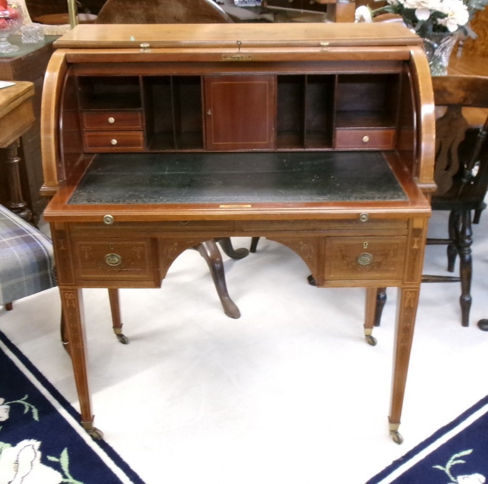 An antique Edwardian inlaid mahogany cylinder top writing desk featuring an open curved roll top, fitted internal compartments, a green leather writing surface, and tapered square legs with brass castors. View IX