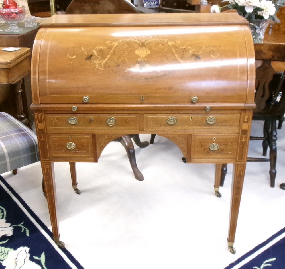 An antique Edwardian inlaid mahogany cylinder top writing desk featuring an open curved roll top, fitted internal compartments, a green leather writing surface, and tapered square legs with brass castors. View X