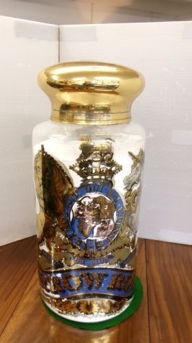 A large clear glass Victorian apothecary jar featuring a gilded coat of arms and arrowroot inscription topped with a fitted domed brass lid.
