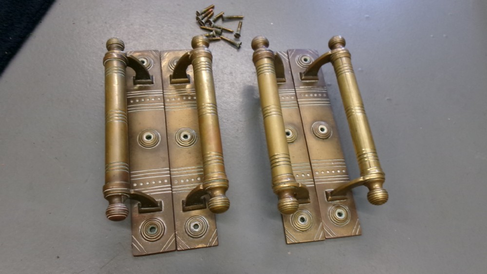 Two pairs of Arts and Crafts brass pull door handles featuring rectangular backplates with incised geometric line detailing and turned cylindrical bar grips with spherical finials.
