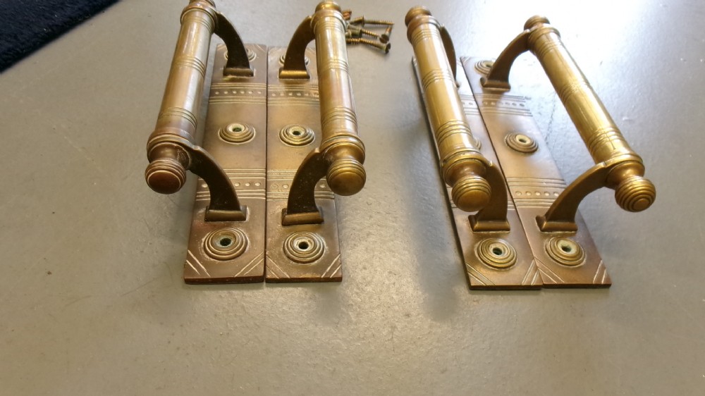 Two pairs of Arts and Crafts brass pull door handles featuring rectangular backplates with incised geometric line detailing and turned cylindrical bar grips with spherical finials. View V