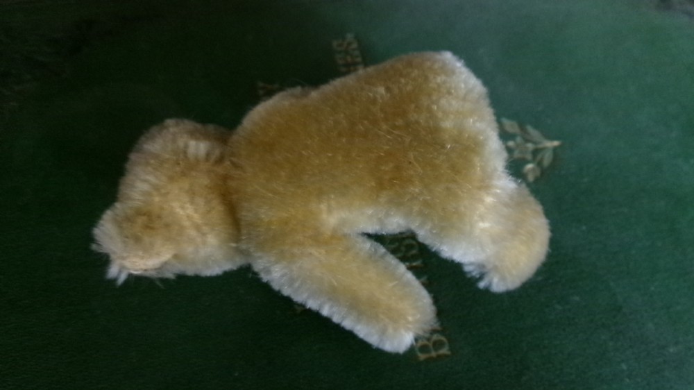 A miniature honey-gold mohair teddy bear sitting down with hand-stitched facial details and a metal button in its left ear. View II