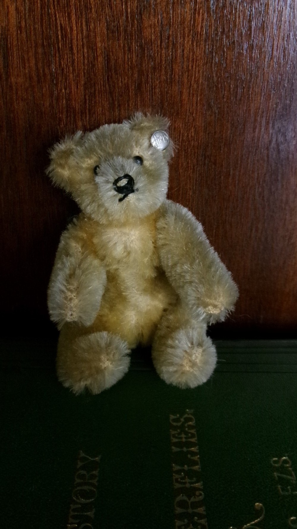 A miniature honey-gold mohair teddy bear sitting down with hand-stitched facial details and a metal button in its left ear.
