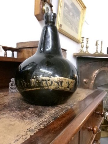A large dark green hand-blown Georgian glass onion bottle with a bulbous body, narrow neck, and a faded gilt label near the base. View V