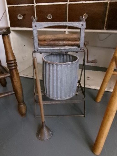 A miniature vintage clothes mangle crafted with a grey iron frame, wooden rollers, a hand crank, an attached galvanised metal tub, and a wooden laundry posser.