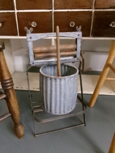 A miniature vintage clothes mangle crafted with a grey iron frame, wooden rollers, a hand crank, an attached galvanised metal tub, and a wooden laundry posser. View II