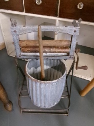 A miniature vintage clothes mangle crafted with a grey iron frame, wooden rollers, a hand crank, an attached galvanised metal tub, and a wooden laundry posser. View III