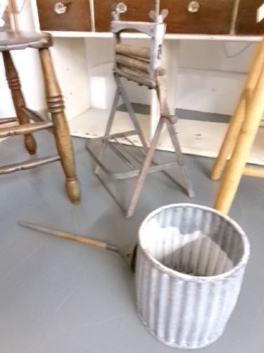 A miniature vintage clothes mangle crafted with a grey iron frame, wooden rollers, a hand crank, an attached galvanised metal tub, and a wooden laundry posser. View V