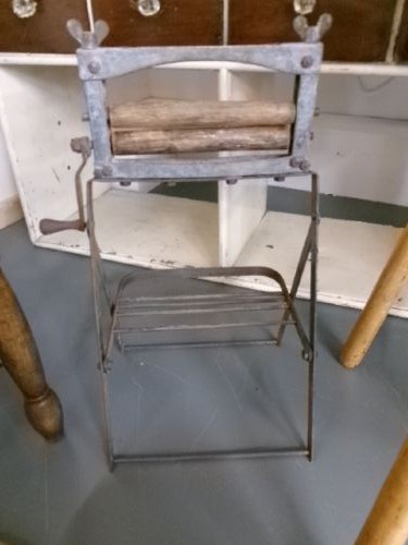 A miniature vintage clothes mangle crafted with a grey iron frame, wooden rollers, a hand crank, an attached galvanised metal tub, and a wooden laundry posser. View VI