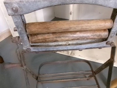 A miniature vintage clothes mangle crafted with a grey iron frame, wooden rollers, a hand crank, an attached galvanised metal tub, and a wooden laundry posser. View VIII