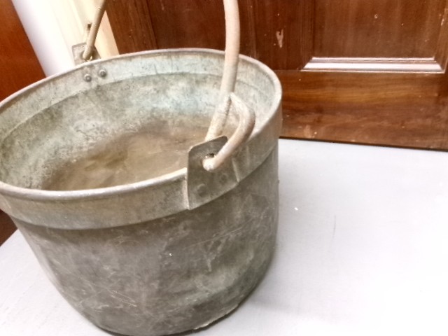 A Victorian metal cauldron featuring a deep circular body, a folded top rim, surface oxidisation, and an arched swing handle attached to side loops. View V