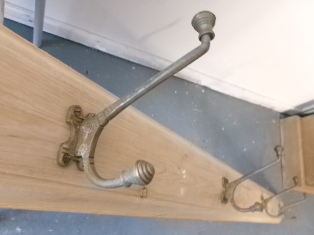 A long vertical pine wood backboard fitted with four double-pronged brass coat hooks. View V