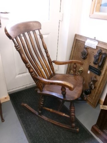 A Victorian dark oak rocking chair featuring a shaped crest rail, vertical spindles, turned arm supports, a solid wooden seat, and curved runners.