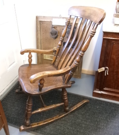 A Victorian dark oak rocking chair featuring a shaped crest rail, vertical spindles, turned arm supports, a solid wooden seat, and curved runners. View III