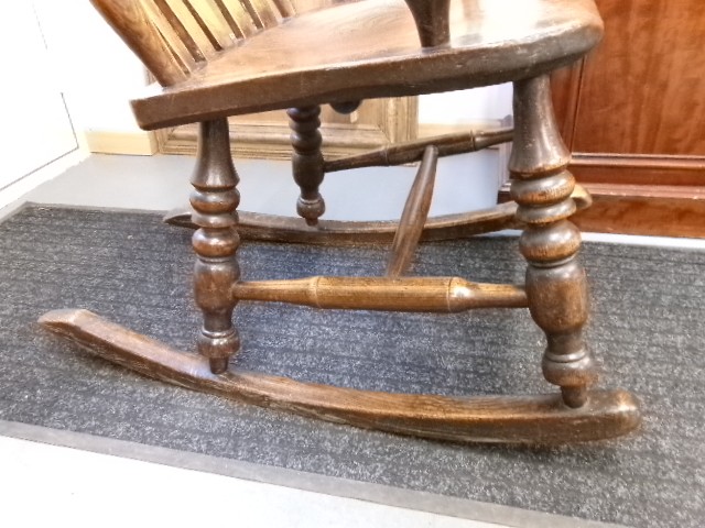 A Victorian dark oak rocking chair featuring a shaped crest rail, vertical spindles, turned arm supports, a solid wooden seat, and curved runners. View V