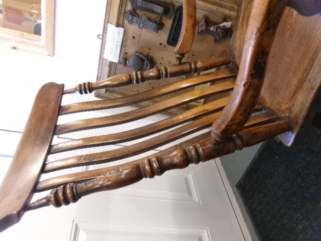 A Victorian dark oak rocking chair featuring a shaped crest rail, vertical spindles, turned arm supports, a solid wooden seat, and curved runners. View VI