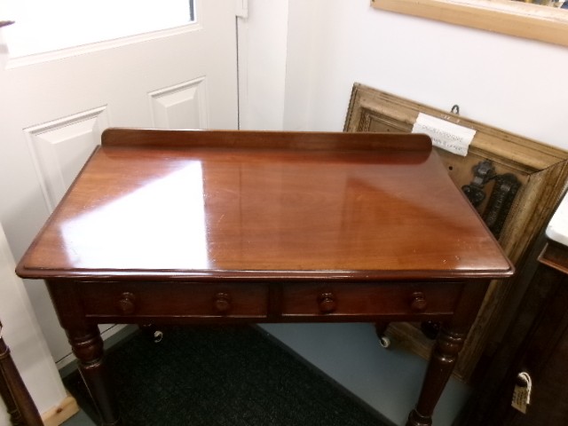 A nineteenth-century Victorian mahogany writing table featuring a low rear gallery, twin drawers with turned wooden knobs, and turned tapered legs resting on white ceramic castors. View II