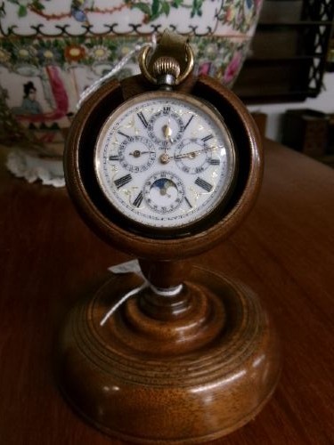 A gunmetal calendar moonphase pocket watch with an enamel dial displayed upright inside a turned wooden holder.