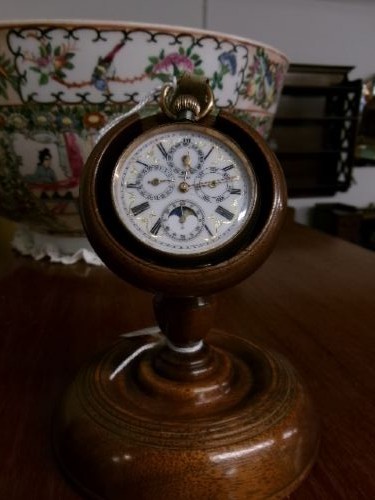A gunmetal calendar moonphase pocket watch with an enamel dial displayed upright inside a turned wooden holder. View IV