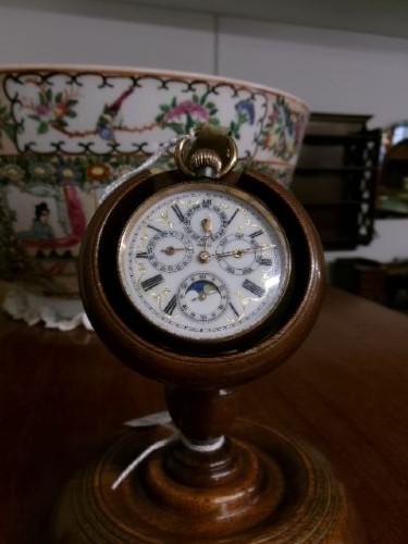 A gunmetal calendar moonphase pocket watch with an enamel dial displayed upright inside a turned wooden holder. View V