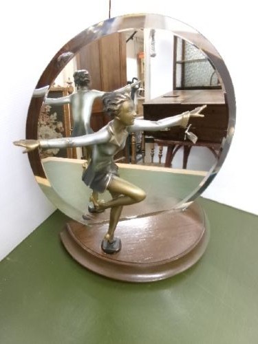 An Art Deco spelter figure of a dancing female posing dynamically in front of a circular bevelled glass mirror set on a wooden plinth base.
