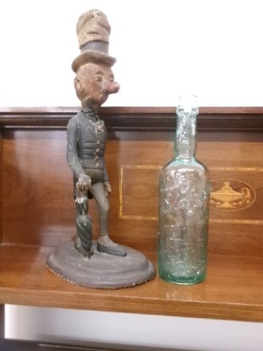 A carved and painted wooden figure of a caricatured man wearing a top hat and tailcoat, holding an umbrella while standing on a rounded grey base. View II