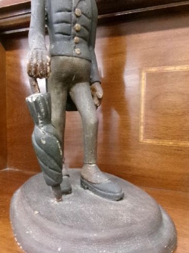 A carved and painted wooden figure of a caricatured man wearing a top hat and tailcoat, holding an umbrella while standing on a rounded grey base. View III