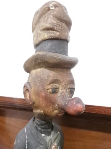 A carved and painted wooden figure of a caricatured man wearing a top hat and tailcoat, holding an umbrella while standing on a rounded grey base. View IV